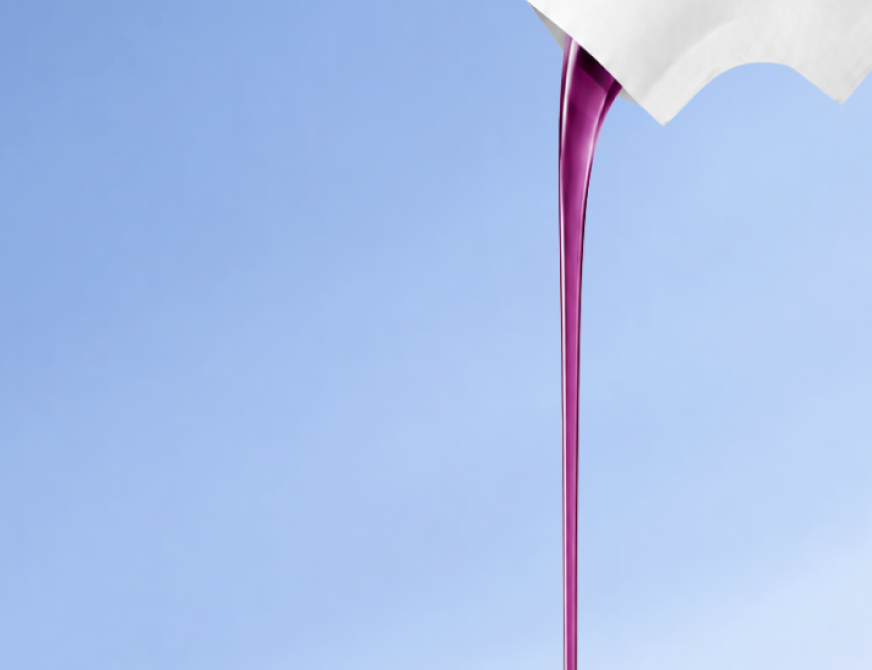A thin ribbon of purple DayShift liquid pouring from a white sachet against a soft blue background.